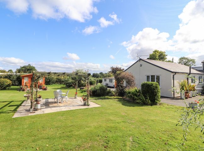 A garden with a white patio table and chairs under a wooden pergola and a small white house in the background at The Flat At Brwyn in Brynford