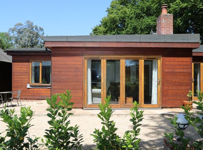 The exterior of a single story wooden house with large glass doors and windows surrounded by green plants at Badgers Sett in Titchfield Common