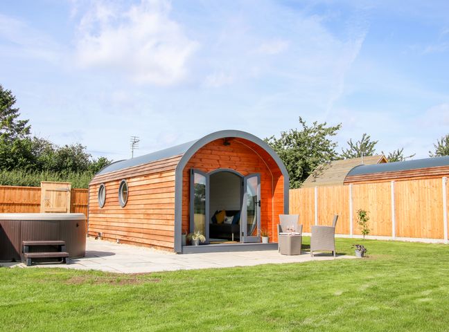 A wooden pod with open doors on a patio with chairs and a hot tub in a fenced garden at Withy Willows in Diddlebury near Ludlow