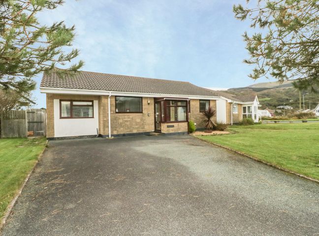 A bungalow with a paved driveway and grass lawn surrounded by trees at Dehangfa in Fairbourne
