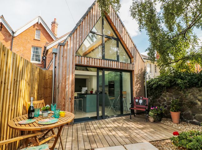 A wooden deck with outdoor table and chairs in front of a modern house with large glass windows at Little Mornington in Malvern