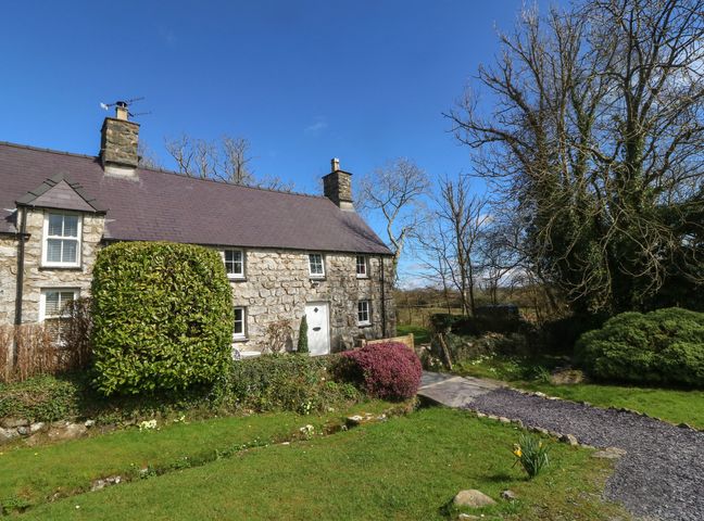A stone cottage with a slate roof surrounded by trees and shrubs with a gravel path and grassy lawn at Y Bwthyn Ty Du in Pwllheli