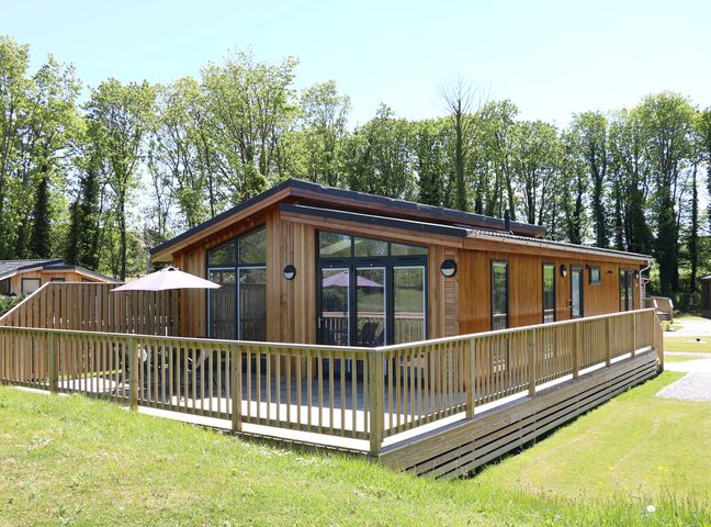 A wooden cabin with large windows and a surrounding wooden deck with a table and umbrella outside in a grassy area