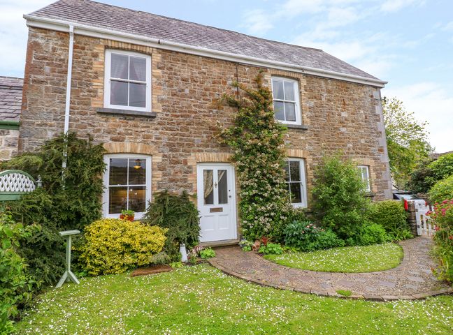 The front of a stone house with a white door and four windows surrounded by green bushes and a stone path leading to the door at Castle School House in Kidwelly