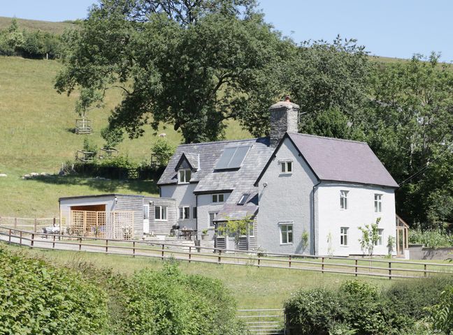A white cottage with a stone chimney and an adjacent wooden structure surrounded by trees and grass fields at Lower Hodre in Chapel Lawn near Bucknell