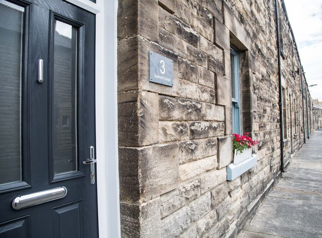 A stone exterior wall with a black door and a window with a flower box at Seashell Cottage in Amble-By-The-Sea