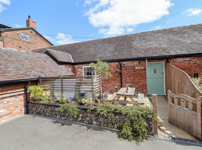 An outdoor patio area with a wooden picnic table a bottle on the table brick buildings with a green door and stone roof at The Dairy in Chester
