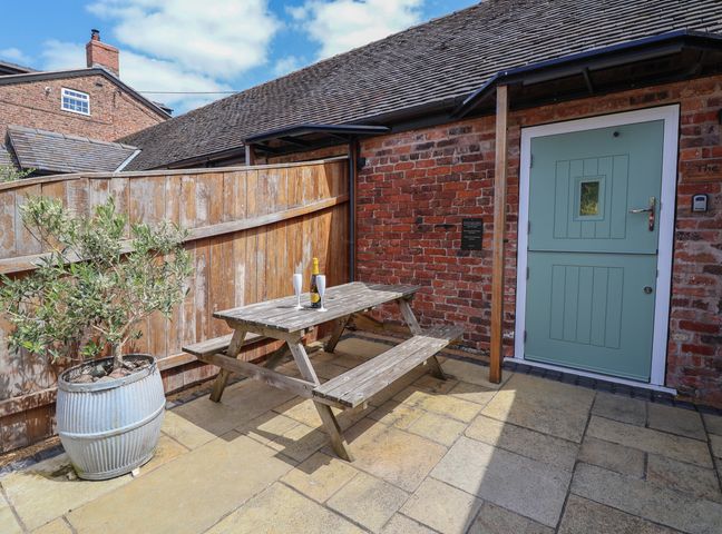 A wooden picnic table with two white wine glasses and a bottle on it a potted plant next to a wooden fence and a blue door on a stone patio at The Parlour in Chester