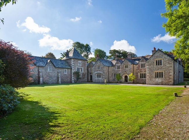 A large stone manor house with multiple chimneys and slate roofs surrounded by green lawn and trees at Great Bidlake Manor in Bridestowe near Lydford
