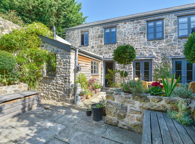 An outdoor stone patio with wooden benches potted plants and a stone planter outside a stone house with multiple windows at Dairy Cottage in Chagford