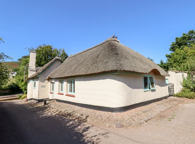 A thatched roof cottage with light walls and blue window frames by a dirt road at Alice Cottage in Bratton near Minehead