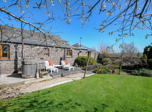 A garden with a grass lawn and stone building with wooden hot tub and patio chairs at Swallow Barn in St Kew