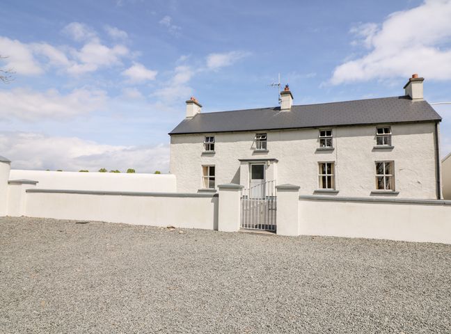 A gravel driveway leading to a two-story white house with a gray roof and a white fence with a metal gate at Grange Farmhouse in Fethard-on-Sea County Wexford