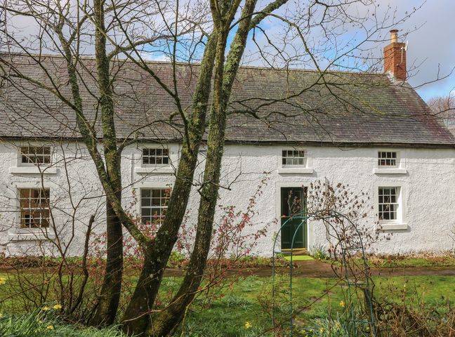 A white cottage with a slate roof and green door seen through trees and garden at The Cottage Polwarth Crofts in Duns