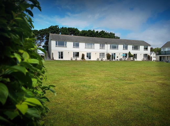 A long white two-story building with several windows and doors overlooking a large grass lawn with trees in the background at Driftwood in Bude