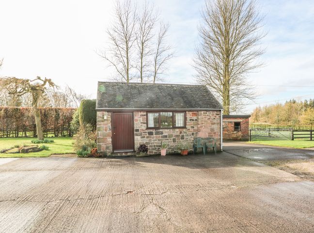 A stone building with a wooden door and window with two plastic chairs outside on a concrete driveway at Cordwainer Cottage in Bagnall