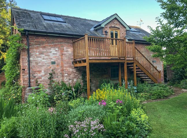 A brick house with a wooden balcony and stairs surrounded by a garden with flowers and plants at The Hayloft in Much Cowarne near Bishop's Frome