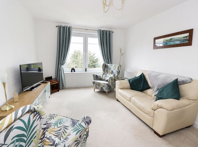 A living room with a beige sofa, two patterned armchairs, a wooden TV stand, a television, and a window with blue curtains at MACS Place in Largs
