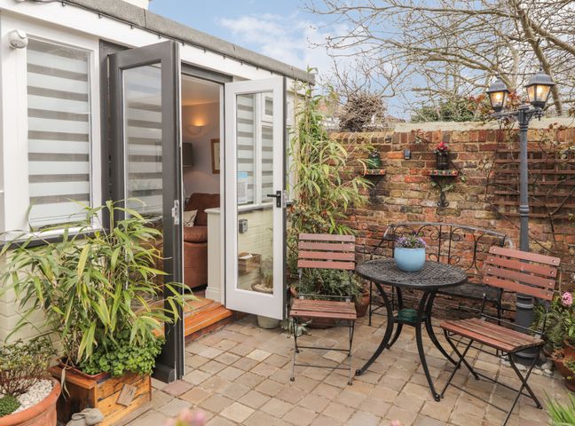 A small outdoor patio with a round metal table and wooden chairs against a brick wall at Rose Cottage in Deal