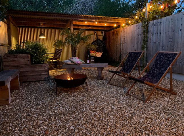 An outdoor seating area with two chairs a wooden bench a fire pit a low table with cushions and wine glasses under string lights at The Old Brewhouse & Piggery in Ningwood near Shalfleet