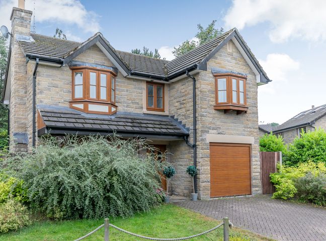 A stone house with wooden framed windows and a garage door surrounded by shrubs and greenery at 2 Carr Farm Close in Glossop