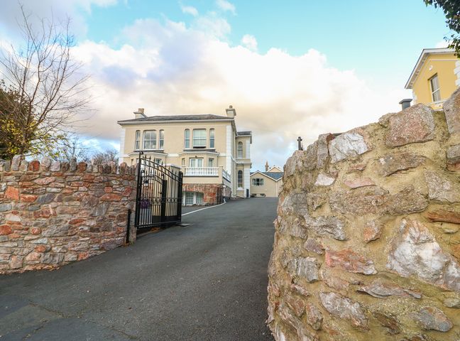 A driveway with a black gate leading to a beige house with multiple windows and stone walls on both sides at Cottage 1 Newcourt in Torquay