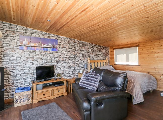 A bedroom with a wooden bed a black leather sofa a TV on a wooden stand and a stone wall at Blackbrae Cabin in Glenboig near Coatbridge Glasgow and Stirling