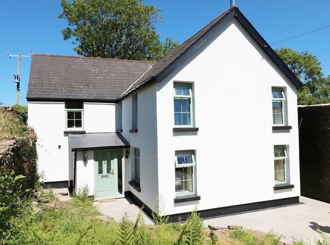A white two-story house with green framed windows and door surrounded by greenery at Ty Ffarm at Gellilwch in Pontypridd