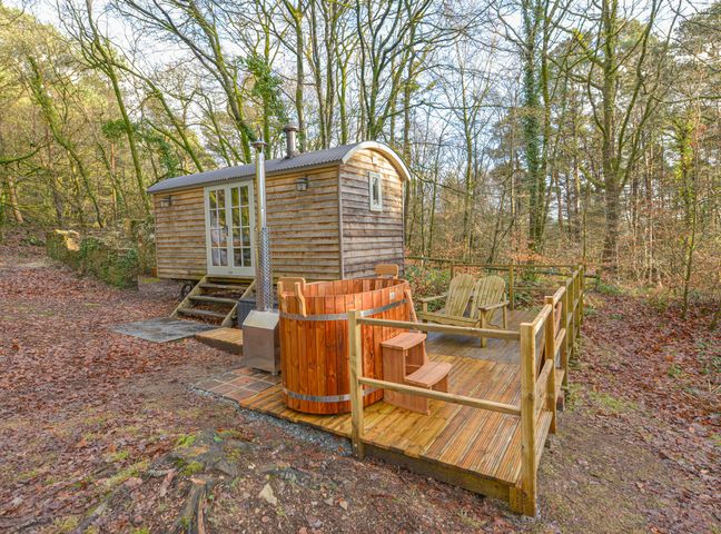 A wooden shepherds hut with a deck featuring two chairs and a wooden hot tub in a forest setting at Rock View Shepherd's Hut in Gulworthy near Tavistock