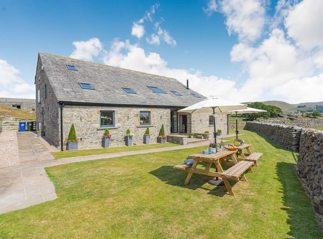 A stone house with a slate roof and outdoor wooden picnic tables with umbrellas in a grassy yard at Dallicar House in Giggleswick near Settle