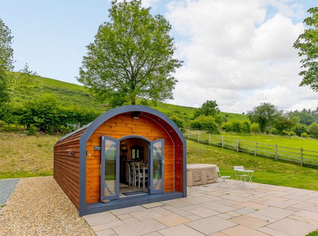 A wooden pod with glass doors open showing a dining area outside a hot tub on a stone patio at Stiperstones Pod in White Grit near Chirbury