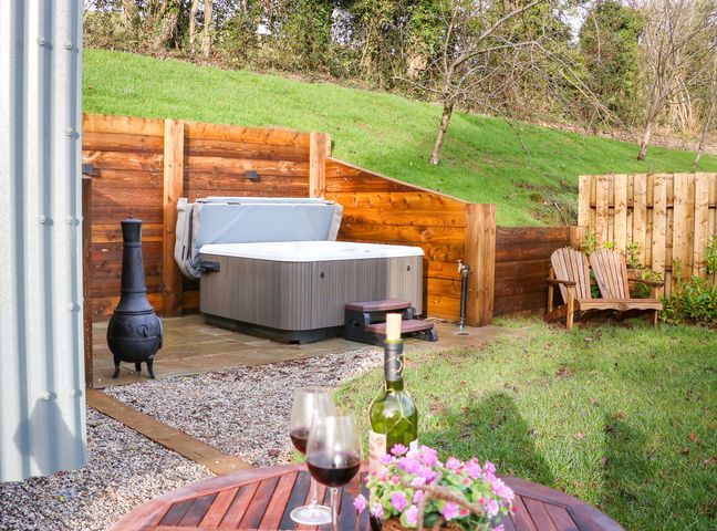 A backyard with a hot tub wooden bench a table with wine glasses and flowers and a metal chiminea at Little Silver Fox in High Bickington