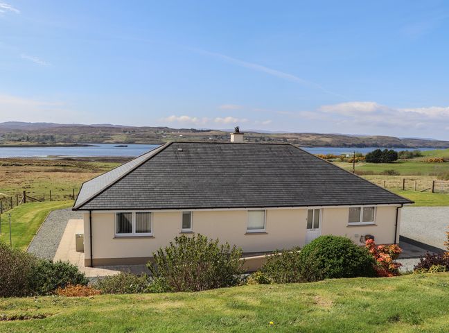A single story house with a dark roof and cream walls surrounded by grass and bushes near a body of water at Burnside Cottage in Portree