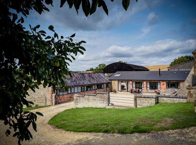 A courtyard with a gravel driveway and stone and brick buildings with a lawn and outdoor seating at Wytons Piece in Chipping Norton