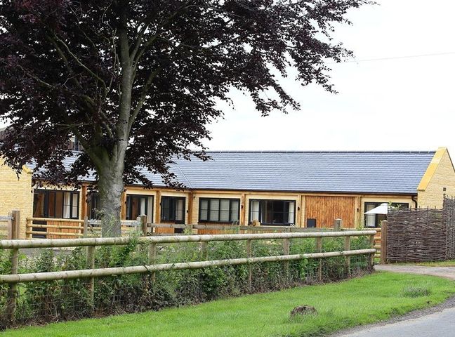 A single-story building with multiple windows behind a wooden fence and a large tree at Park Stables Wyck Beacon near Bourton-on-the-Water