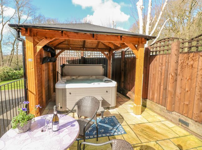 An outdoor patio with a hot tub under a wooden gazebo and a table with chairs and a bottle at Bluebell Cottage Alfreton near Oakerthorpe
