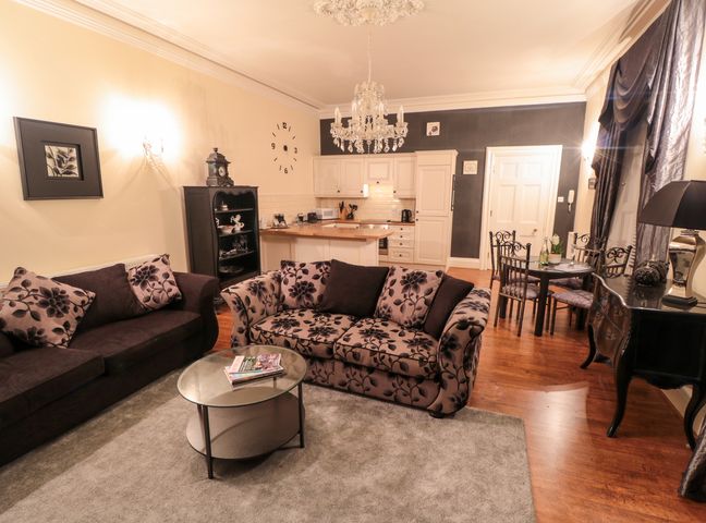 A living room with patterned sofas a glass coffee table a chandelier a dining table and kitchen area at Sneaton Hall Apartment 4 in Sneaton