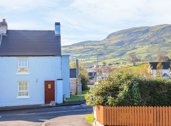 A street with a blue house with a brown door and windows next to a hedge with a wooden fence and hills in the background at Cranny Corner in Carnlough