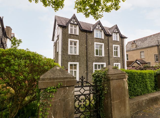 A three-story stone house with white framed windows behind a wrought iron gate and stone pillars in Criccieth