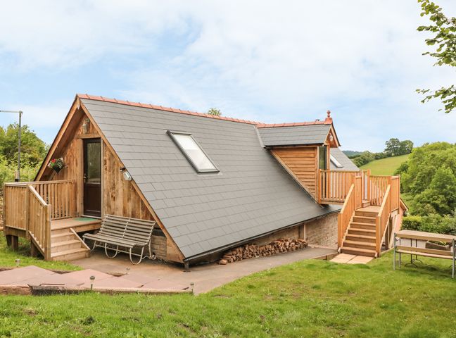 A wooden house with a grey sloped roof and stairs with railings surrounded by grass and trees at The Lookout in Llangrove