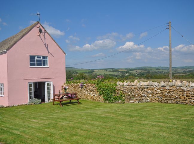 A pink house with open double doors and a picnic table on a grass lawn with a stone wall and countryside beyond at Cliff Farm No 2 Cottage Burton Bradstock