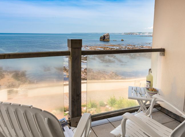 A balcony with two white chairs a small table with glasses and a bottle overlooking the sea at 3 Thurlestone Rock in Thurlestone
