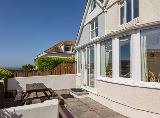 An outdoor patio area with a wooden picnic table and white wall next to a house with large windows at Cottage View in Hope Cove