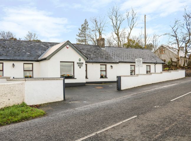 A white single-story cottage with several windows and flower boxes behind a low wall on a paved road at Woodleigh Cottage in Ballycastle