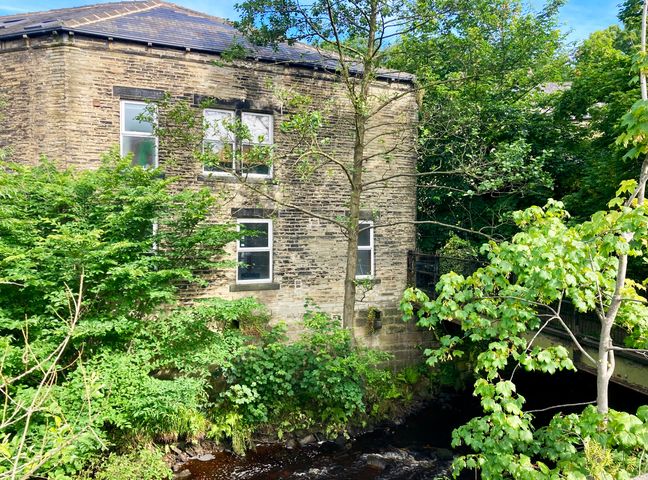 A stone building next to a small river surrounded by green trees and a metal bridge at The Old Riverside School House in Hebden Bridge