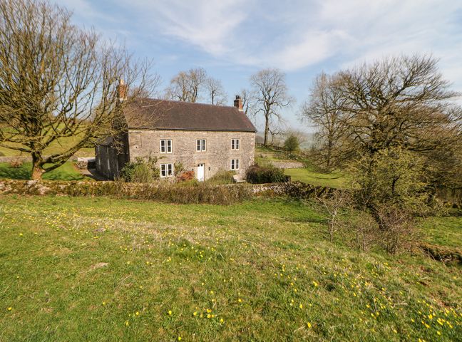 A stone house with multiple windows and a front door surrounded by grassy land and trees at Slade House in Ilam Staffordshire