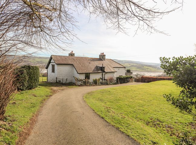 A single-story white cottage with a tiled roof at the end of a curved driveway with grass and trees around at Cymryd Uchaf in Conwy