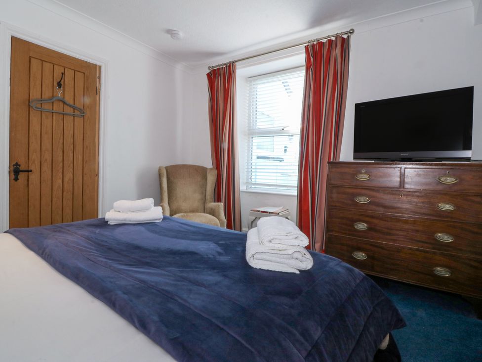A bedroom with a bed covered in a blue blanket with folded towels on top an armchair by a window with red striped curtains a wooden dresser with a television on top and a wooden door Bay View Apartment Porthmadog