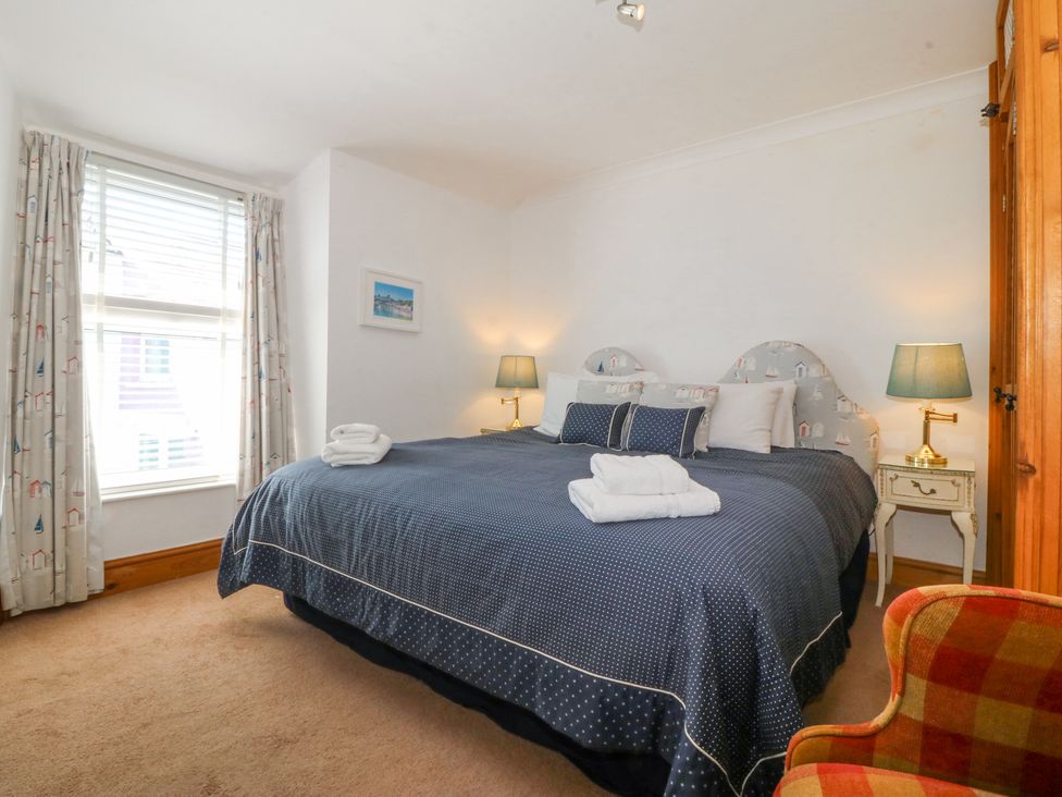 A bedroom with a large bed with blue bedding two bedside tables with lamps a window with curtains and a checkered armchair at Bay View Apartment in Porthmadog