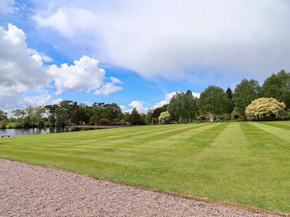 A lawn next to a lake with trees and a blue sky at South Tower Cottage in Capesthorne near Macclesfield